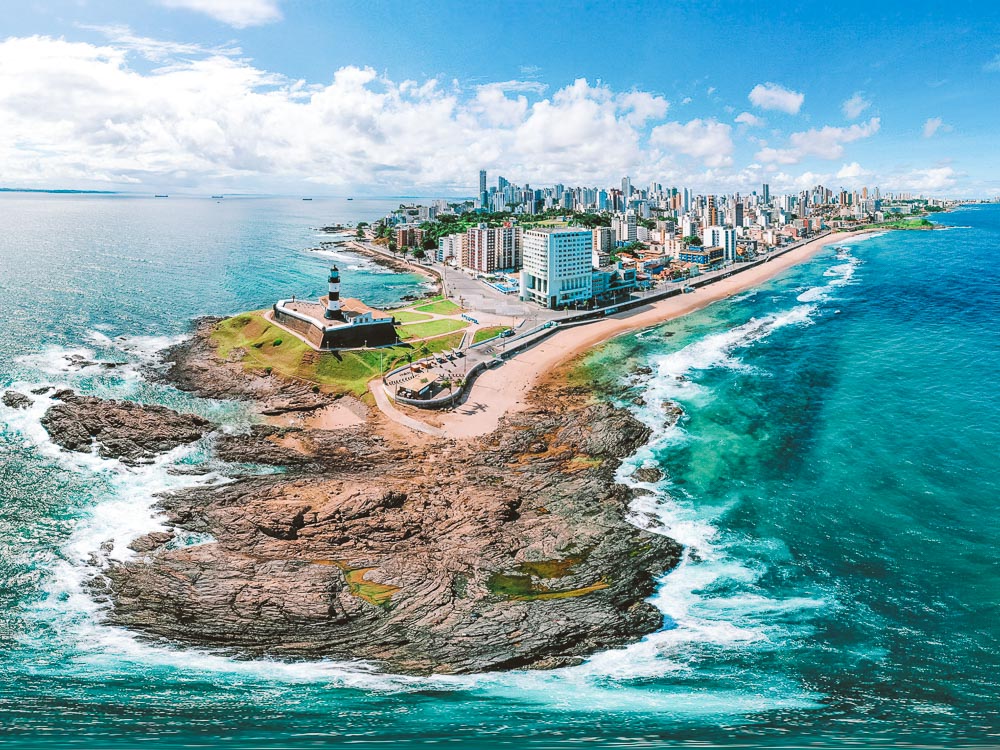 Vista aérea de Salvador na Bahia, na imagem mostra uma ilha com areia ao redor, e a cidade atrás com prédios. Uma estrada de chão é possível ver na lateral dos prédios indo de encontro com a cidade.