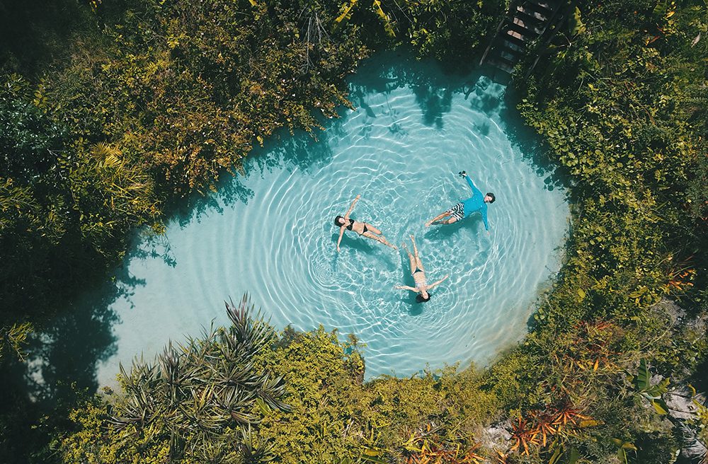 Três pessoas flutuando no fervedouro Por Enquanto, com formato semelhante a uma gota e com água azul cristalina